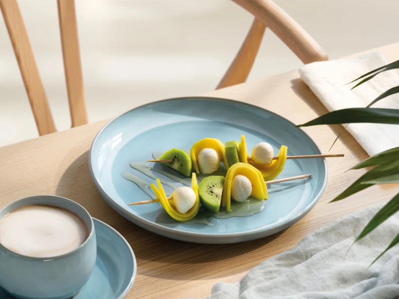 A blue perlemor aqua plate with fruit and mozzarella skewers A blue perlemor aqua plate with fruit and mozzarella skewers next to a cup of coffee on a light wooden table.