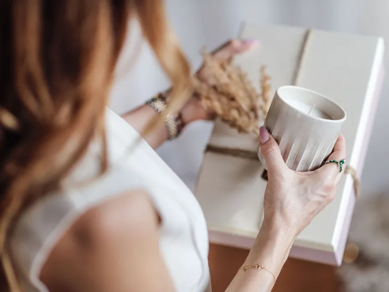 A person holding a Perlemor scented candle in their left hand and a wrapped gift decorated with dried plants.