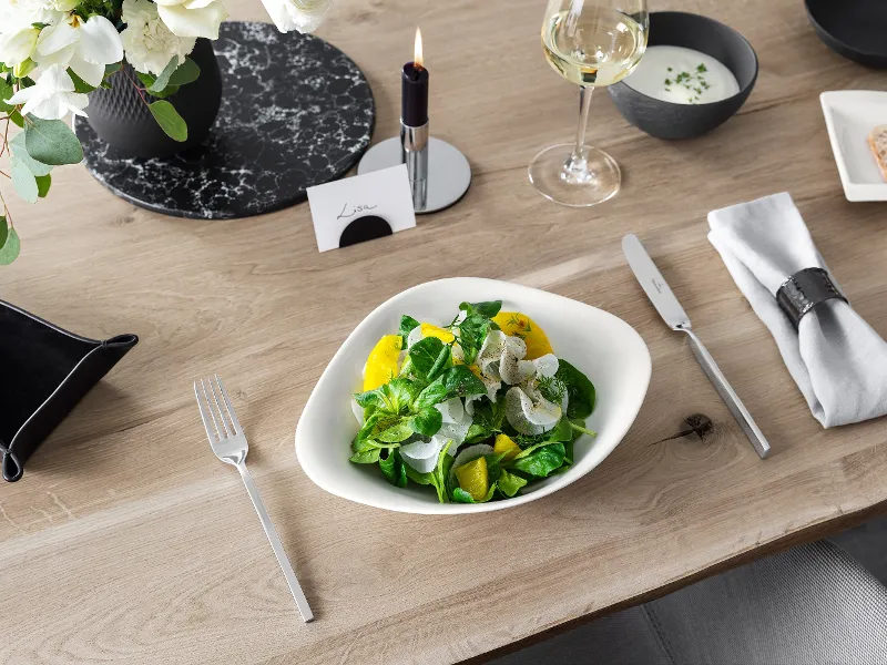 A Villeroy & Boch Vapiano bowl with salad on a wooden table, beside a NewWave knife and fork.