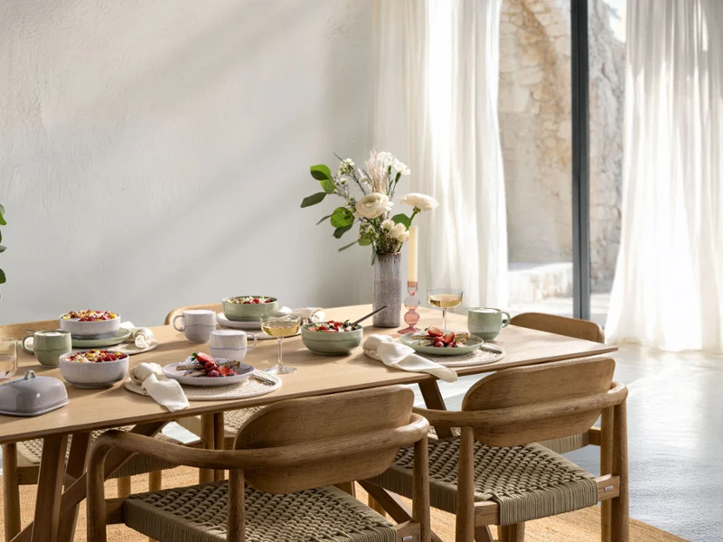 A breakfast table set for six with bowls, plates and flower vases in a sun-filled room with large windows and light-coloured curtains.