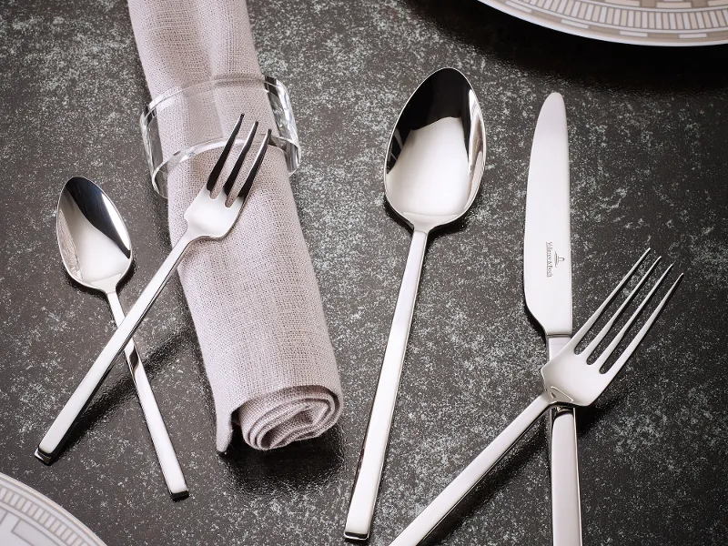 Une présentation de table avec des couteaux La Classica, des fourchettes, des cuillères et une serviette avec un rond de serviette sur une surface structurée foncée. Des assiettes blanches avec des motifs géométriques sont partiellement visibles.