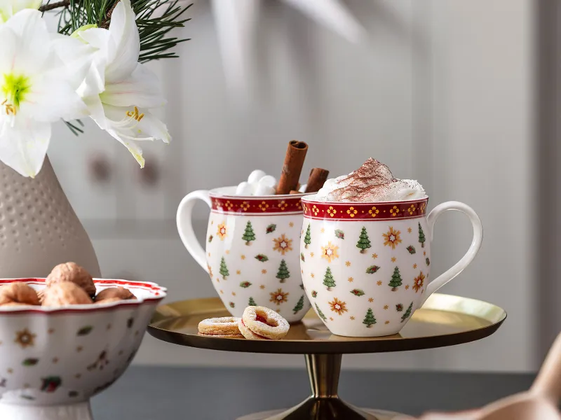 Two festive Toy's Delight cups with whipped cream and cinnamon sticks on a tray, with biscuits and a flower vase next to them.