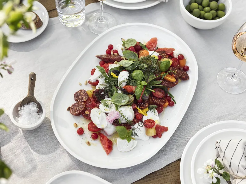 On a white tablecloth, there is a Villeroy & Boch Artesano platter with a mixed salad of tomatoes, herbs and cold cuts. Next to it are bowls of olives, a glass of rosé and a bowl of salt.