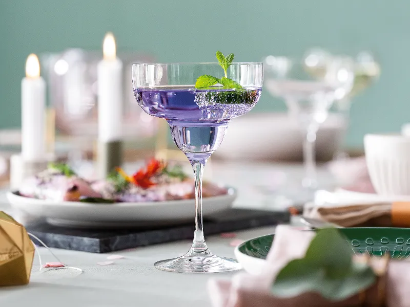 A purple cocktail, garnished with mint in a Purismo margarita glass on a laid table, surrounded by plates, candles and decoration objects.