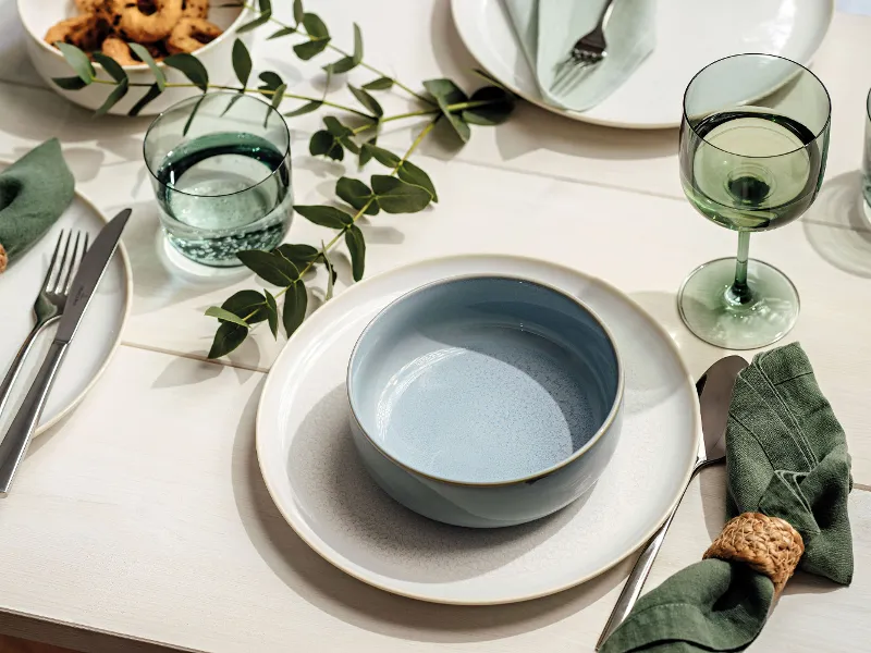 A beautifully set table with a light blue bowl from Villeroy & Boch Crafted on a white plate, green glasses, eucalyptus branches, and green cloth napkins with rings. Other plates, flatware, and food are partially visible.