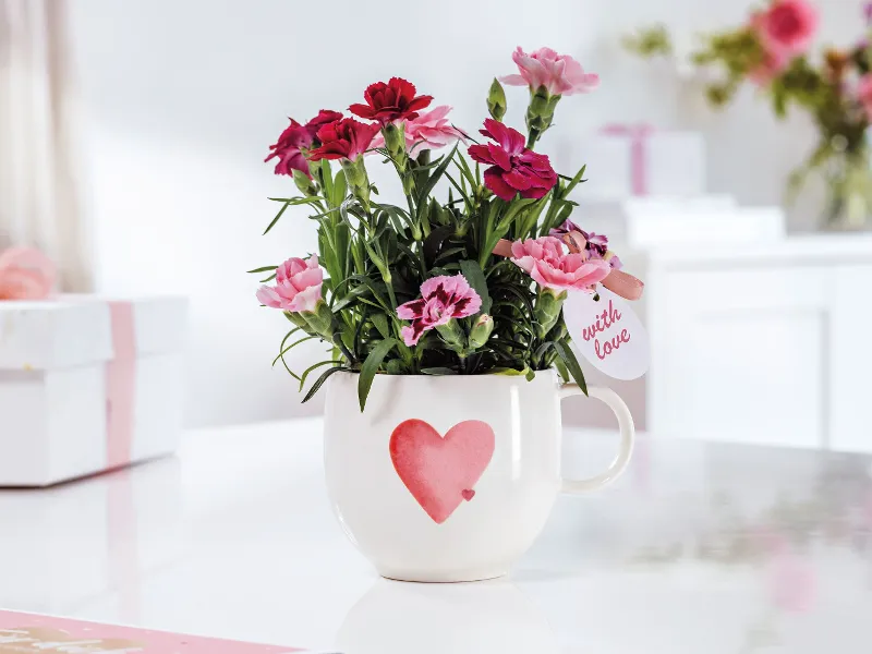 A white With Love cup with a red heart and pink and red flowers on a white table.