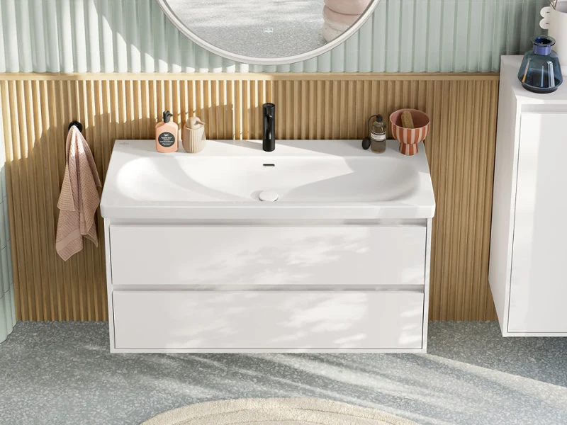 Lavabo espacioso y moderno en colores pastel visto desde arriba, que consta de un mueble de lavabo con mueble bajo blanco, un espejo redondo iluminado, un mueble lateral y un grifo negro de la colección de baño Skyla.