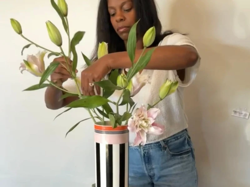 Floral arranging with Memphis Vase Woman carefully arranges green lilies and pale pink flowers in a striped vase, surrounded by coral blooms, creating a calm floral display.
