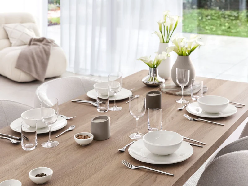 Minimalist Dinner Table with Manufacture Collier Blanc Vases A bright, modern dining table set with white plates, bowls, glassware, grey candles, and vases filled with white calla lilies, in front of large windows overlooking a green garden.