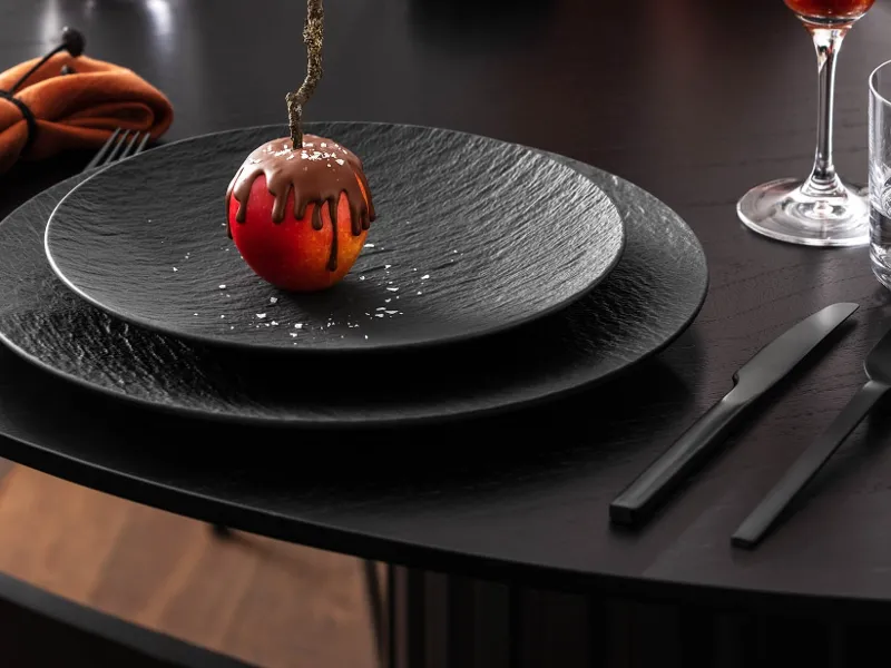 An artistic dessert resembling an apple with chocolate icing, served on an elegant black Manufacture plate, accompanied by modern black Manufacture flatware and glassware.