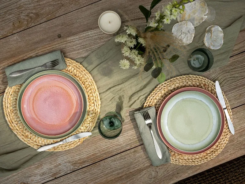 Two Perlemor place settings on a wooden table with round woven placemats, cutlery, a candle, flowers and green plants.