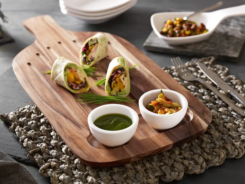 Villeroy & Boch Artesano wooden board with vegetable wraps, green chutney and two small bowls of mixed salsa. Served with cutlery on a woven base, with more plates and a container visible in the background.