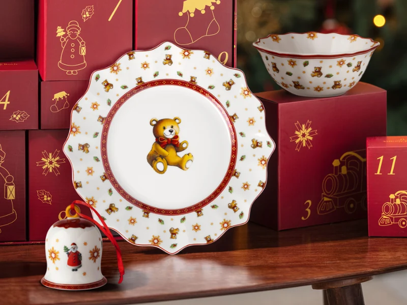 A white plate, a bowl, and a bell with teddy bear and ribbon motifs, displayed in front of red Christmas gift boxes from the Villeroy & Boch Advent calendar.