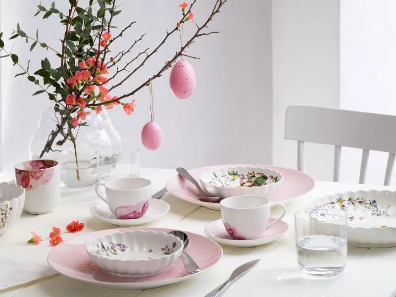 A bright, springtime dining table set with pastel-colored dinnerware from the Rose Garden and Spring Fantasy collections, decorated with floral patterns and Easter eggs hanging from a branch as a centerpiece.