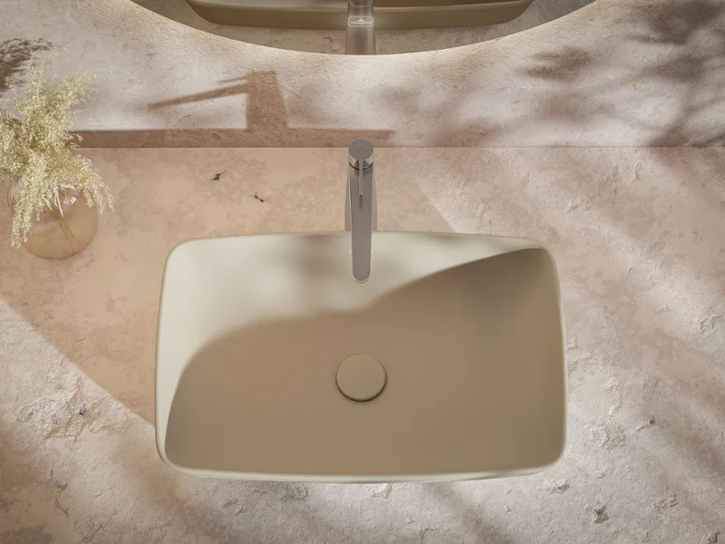 A rectangular beige washbasin with a modern faucet on a stone countertop; next to it is a vase with small white flowers.