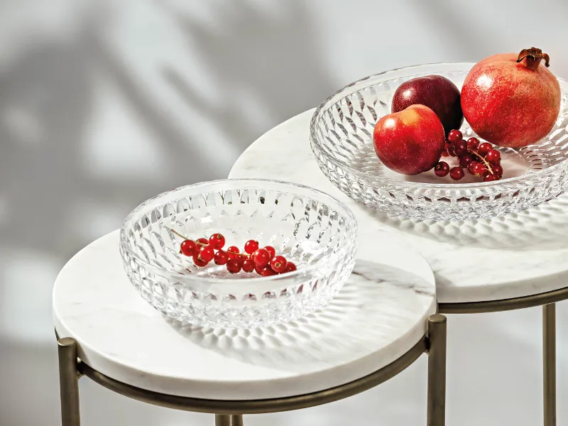 Two clear Boston glass bowls on marble tables with fruit. One contains pomegranates and an apple, the other contains redcurrants.