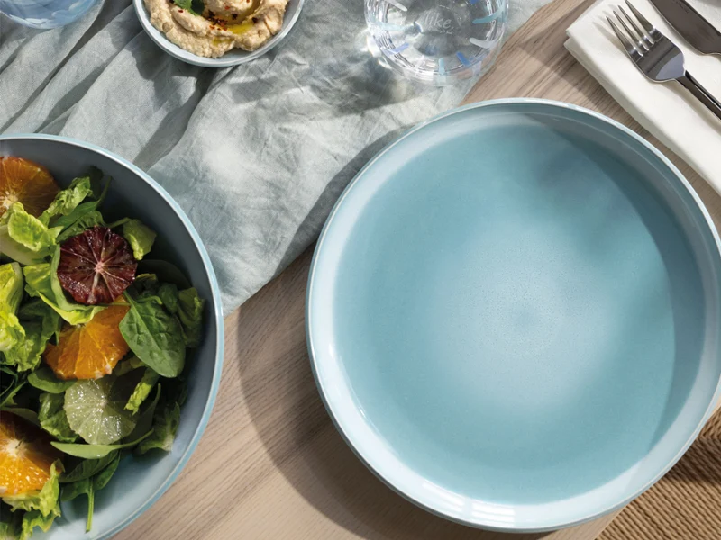 Ein leerer blauer Perlemor Aqua Teller steht neben einer Schüssel mit Salat, Besteck und einer Serviette auf einem Holztisch.
