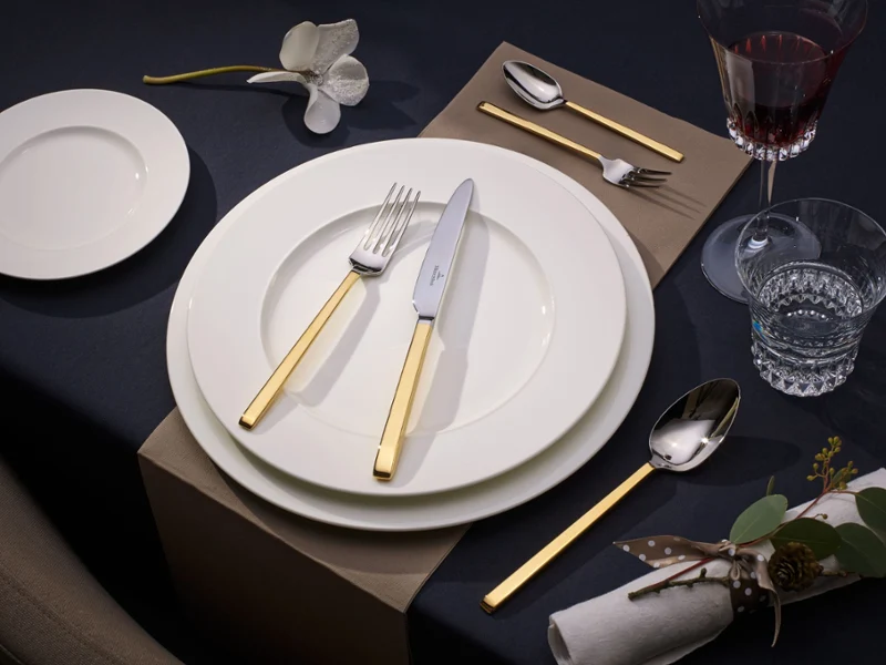 Elegant table setting with gold-handled cutlery on white plates. A wine glass, water glass, brown napkin with silverware, and floral decor enhance the scene.