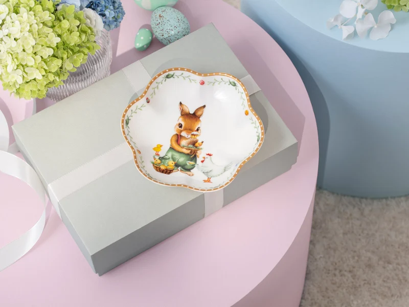 A gift box with a ribbon sits on a pink table, topped with a decorative Annual Easter Edition 2026 plate featuring a rabbit and flowers.
