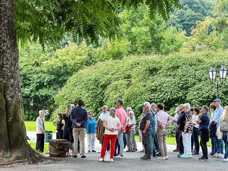 A group of people is standing outdoors near a large tree and a lamppost in the middle of the Villeroy & Boch Park, surrounded by greenery.