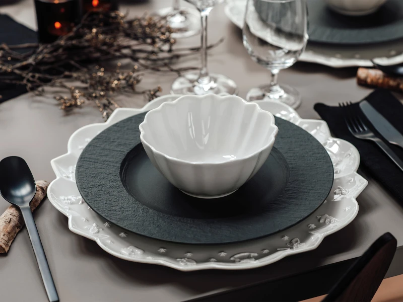 A formal table setting with a Manufacture Plate on top of a Toy´s Delight Royale Classic plate A formal table setting with a white Toy´s Delight Royale Classic bowl on black and white plates, glassware, and black cutlery on a grey table.