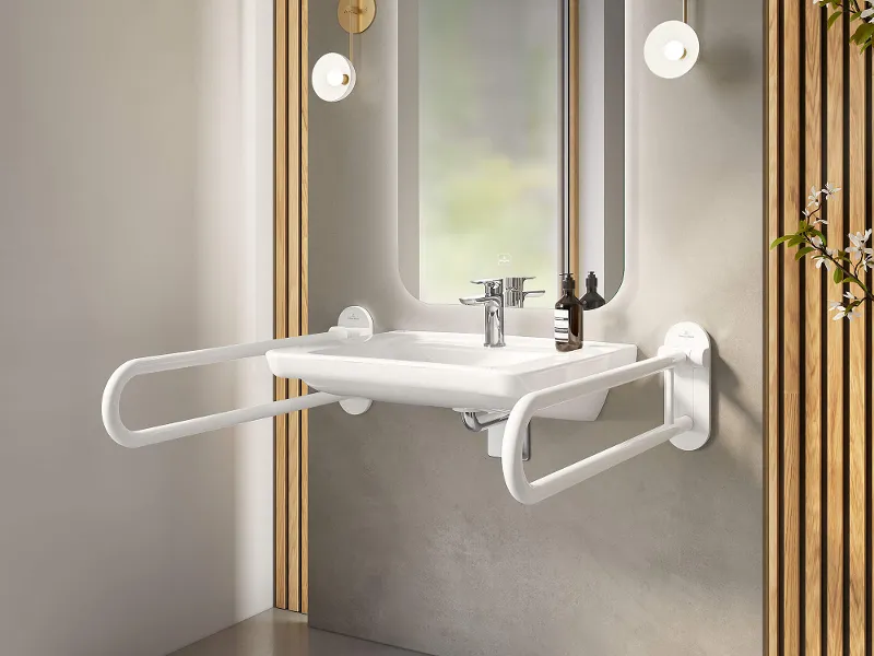Un lavabo ViCare con maniglie pieghevoli e uno specchio con illuminazione d'ambiente su una parete intonacata di grigio.
