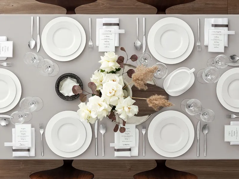 Table setting with Villeroy & Boch Royal white plates, stylish glassware and elegant silver cutlery on a beige tablecloth. The centrepiece features white roses with brown accents. A small menu and a napkin are at each setting.