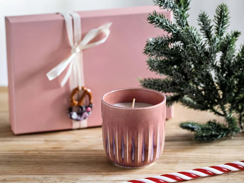 A pink Perlemor scented candle on a wooden surface next to a small fir sprig, a pink gift box with a white bow and ornament and a large striped sugar cane.