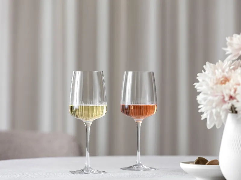 Two Villeroy & Boch Afina wine glasses, one filled with white wine and the other with rosé, are placed on a table next to a vase with pink flowers.