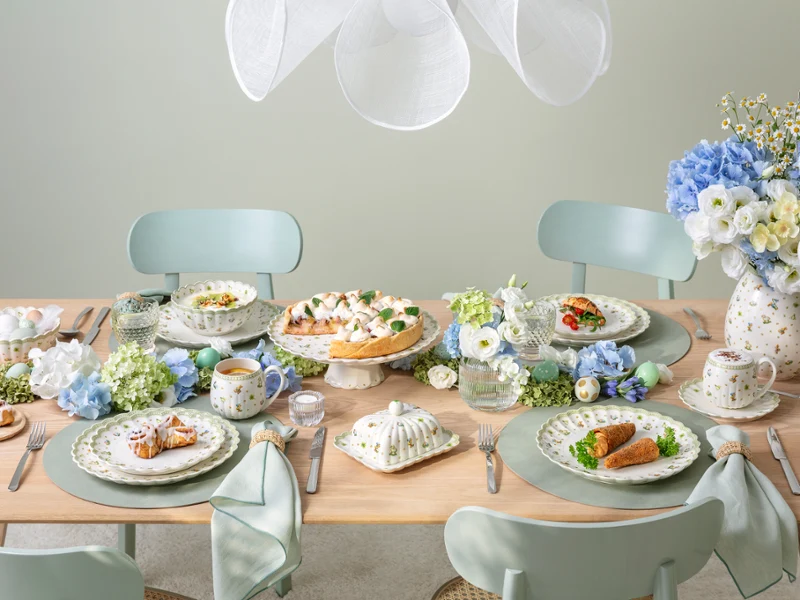 A pastel-themed dining table set for four, featuring plates of food, teacups, blue and white floral arrangements, and elegant tableware, creating a fresh and inviting atmosphere.