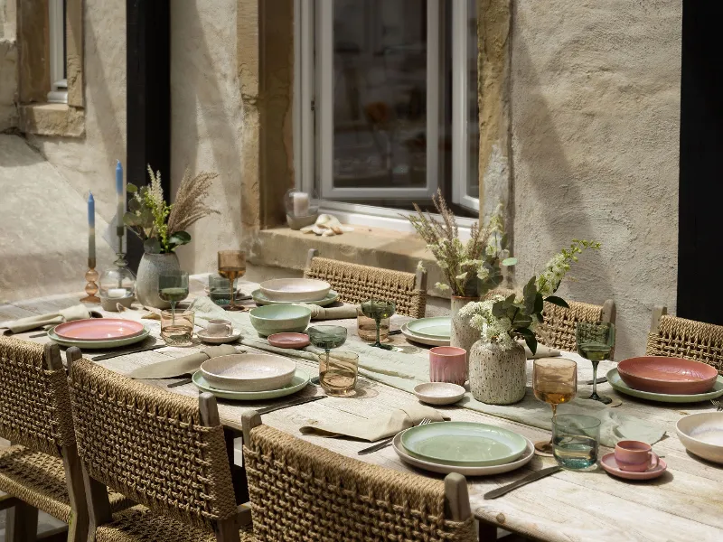 A rustic outdoor dining table setting with woven chairs, like. by Villeroy & Boch pastel-coloured Perlemor plates, glasses and floral centrepieces against the backdrop of a stone wall with a window.