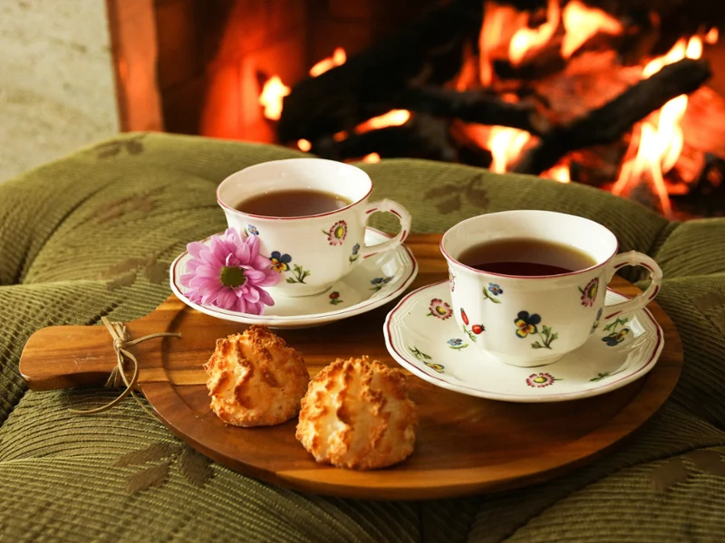 Two floral teacups and saucers on a wooden platter with cookies in front of fireplace