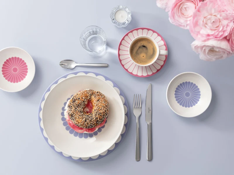 A breakfast set with bagel, cup of coffee, decorative plates, cutlery and flowers on a light blue background.