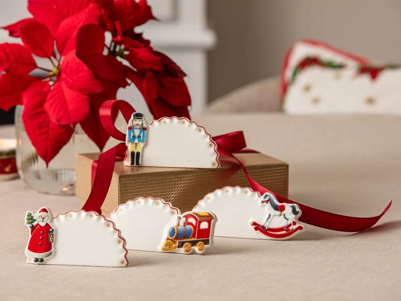 Festive place cards decorated with Christmas figures A table with festive place cards decorated with Christmas figures, a red ribbon and poinsettias in the background.