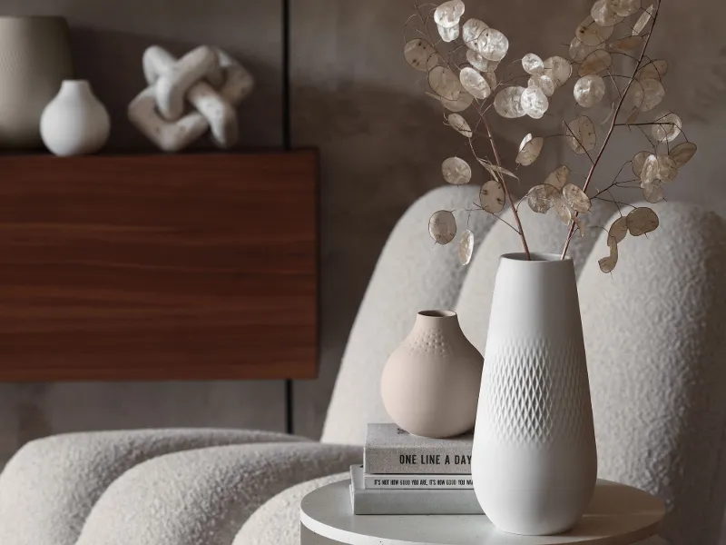 A modern table with a white Manufacture vase with dried flowers, books and decorative objects next to a soft chair in neutral colours in a modern interior.