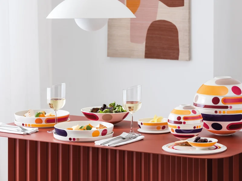 A modern dining table set with the Miami La Boule A modern dining table set with colorful patterned dishes, bowls of food, wine glasses, and abstract wall art in the background.