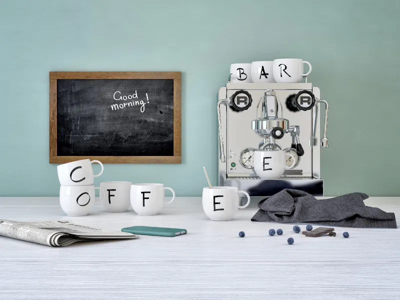 Espresso machine with Villeroy & Boch "Letters" cups spelling out the word "COFFEE" on a table, next to a phone, a newspaper, blueberries and a blackboard with the words "Good morning!".