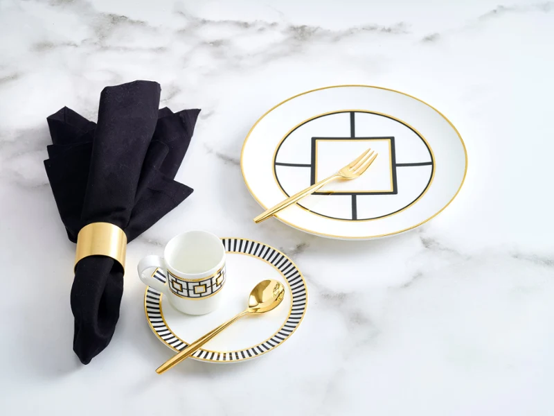 The Metrochic Cafe 4pcs gift set includes a plate, cup, and two pieces of cutlery, all featuring gold accents and a black line design on white porcelain, complemented with a black linen napkin on a white marble table.