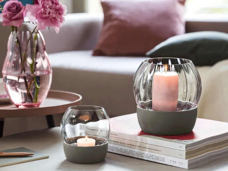 A cosy living room with a pink sofa, a Rose Garden glass vase with flowers, lit candles in Rose Garden glass holders, coffee and books on a round table.