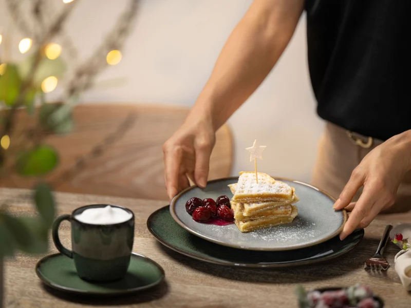 A cozy breakfast scene featuring a Villeroy & Boch Lave Vert dinner plate and cappuccino cup with saucer, paired with a beige Lave breakfast plate holding a beautifully served waffle dish. A woman is reaching out to serve the plate, surrounded by warm lighting and festive, homey holiday decor.