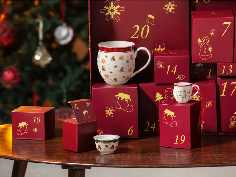 On a wooden table next to a decorated Christmas tree stands a red advent calendar with numbered compartments, cups and bowls from Villeroy & Boch.