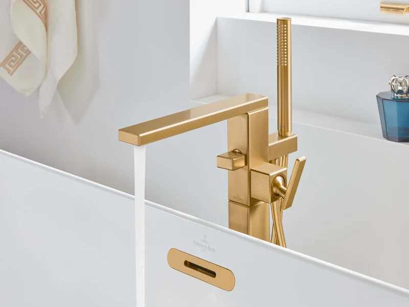 Złota bateria wannowa Mettlach ze słuchawką prysznicową Nowoczesna łazienka z kranem w złotym kolorze, z którego płynie woda, z ręcznikiem wiszącym na ścianie i przedmiotami dekoracyjnymi na półce.