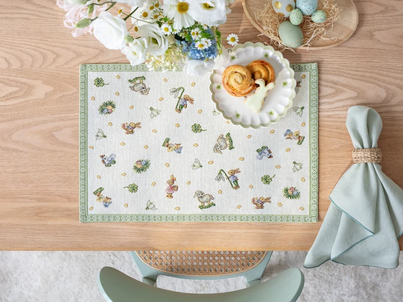 A wooden table set with a Easter Delight placemat A wooden table set with a Easter Delight placemat, pastries on a plate, flowers, dyed eggs, and a folded napkin.