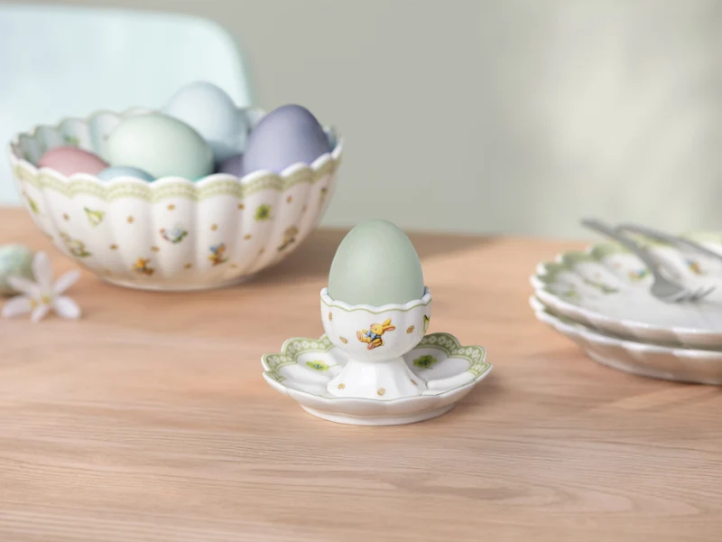 Easter Delight egg cup with Easter decorations A pastel-colored egg in an Easter Delight egg cup with Easter decorations stands on a wooden table with a bowl full of eggs and a plate with cutlery in the background.