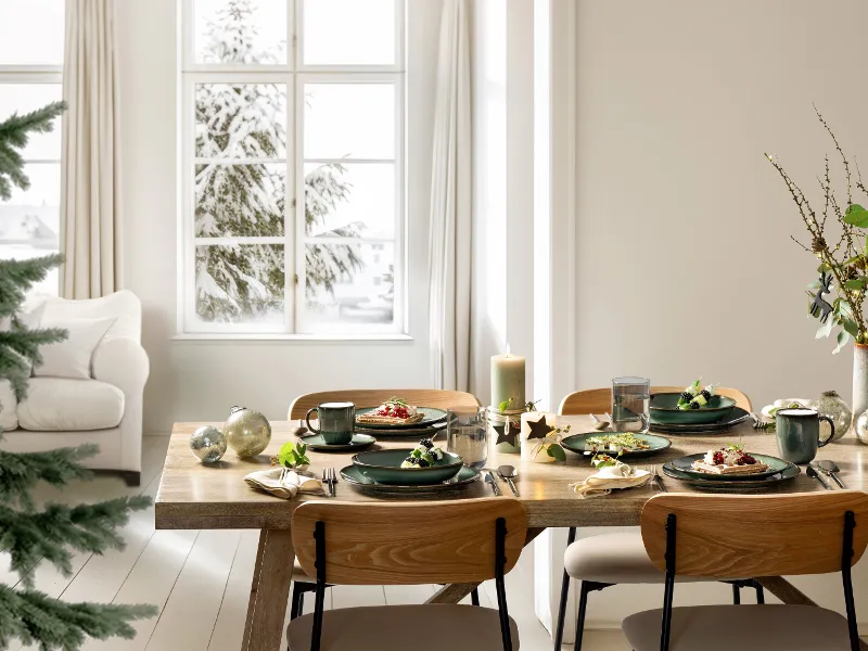 A wooden dining table for four with Lave Vert plates, cups, and candles in a bright room overlooking the snow outside.