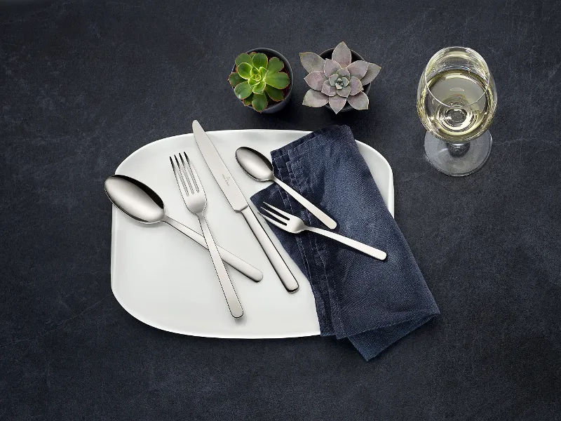 Table setting with a white plate, stainless steel cutlery from the Louis collection, a dark napkin, two small succulents and a glass of white wine on a dark surface.