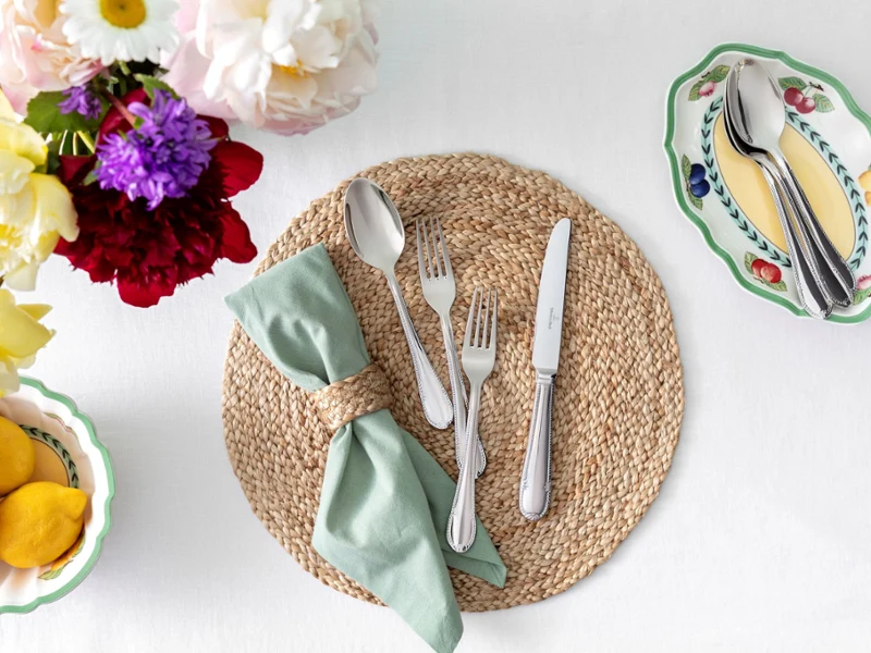 A table setting featuring a jute mat, a bowl of flowers, and a knife and fork arranged neatly.
