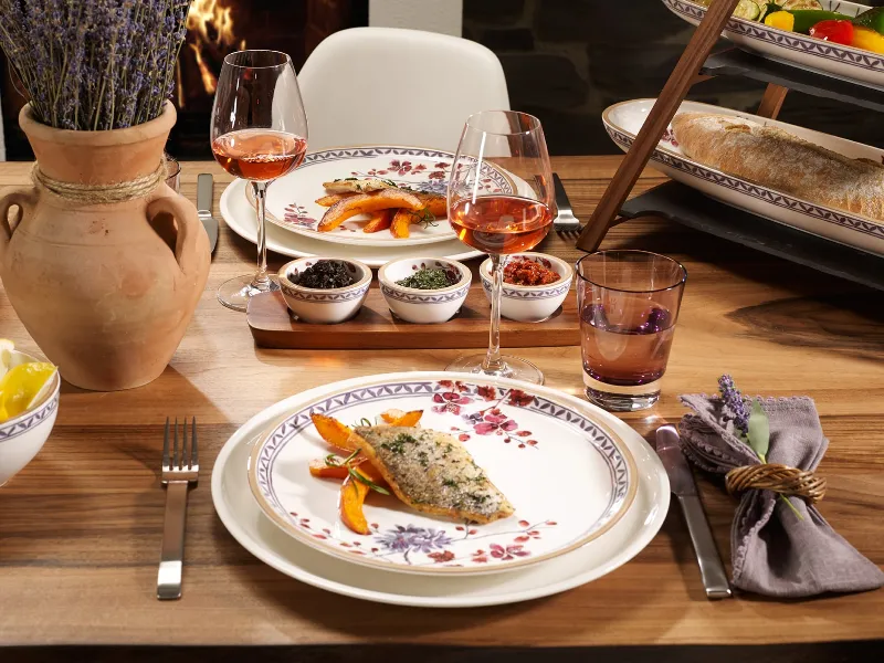 Tavola imbandita per due persone con pesce su piatto Artesano Provencal Lavanda di Villeroy & Boch, carote su piatti da fiori, bicchieri da vino, erbe aromatiche in ciotole, pane e un bouquet di lavanda in un vaso.