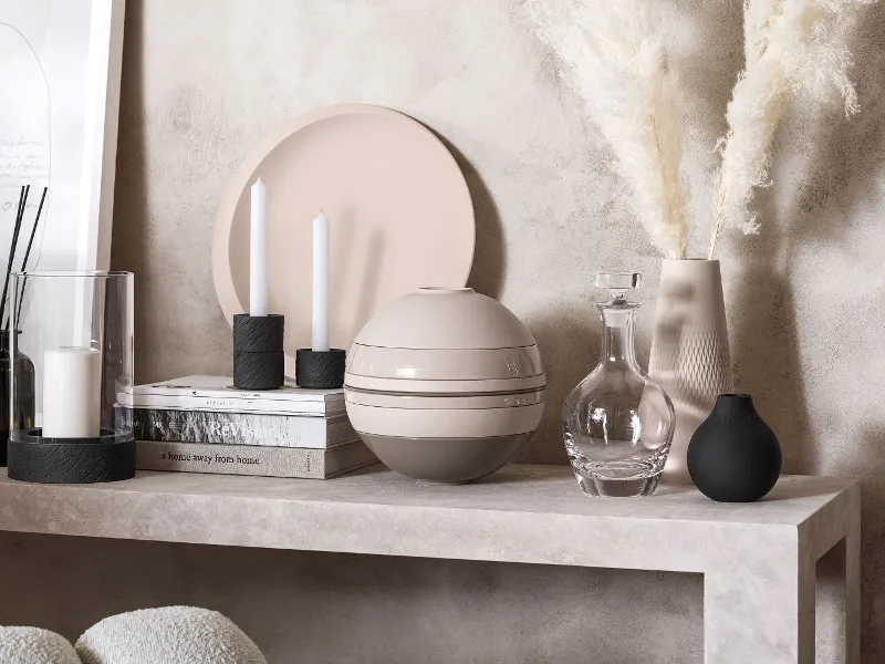 A modern table setting with decorative objects in neutral tones, including candles, a transparent carafe, and a vase with pampas grass and a modern La Boule, in front of a wall with a light texture.