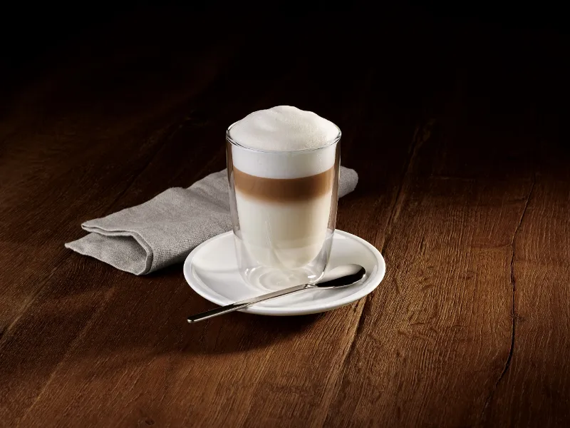 A glass of layered latte with foam on a Villeroy & Boch Coffee Passion saucer, together with a spoon and next to a folded grey napkin on a wooden surface.